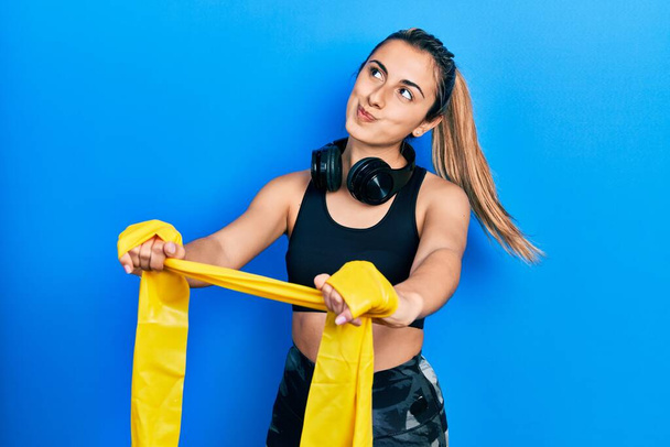 Beautiful hispanic woman training arm resistance with elastic arm band smiling looking to the side and staring away thinking.  - Photo, Image