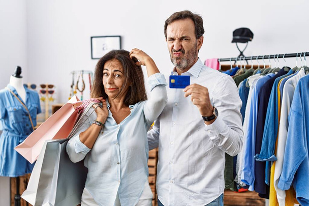 Hispanic middle age couple holding shopping bags and credit card confuse and wondering about question. uncertain with doubt, thinking with hand on head. pensive concept.  - Foto, Imagem