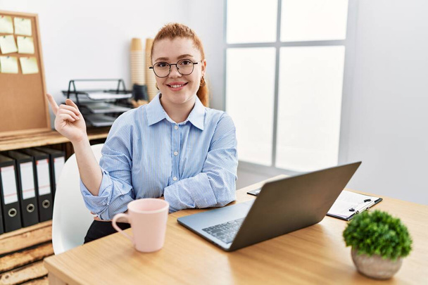 Fiatal vörös hajú nő dolgozik az irodában számítógépes laptop egy nagy mosollyal az arcán, mutató kéz ujjával oldalra nézi a kamerát.  - Fotó, kép