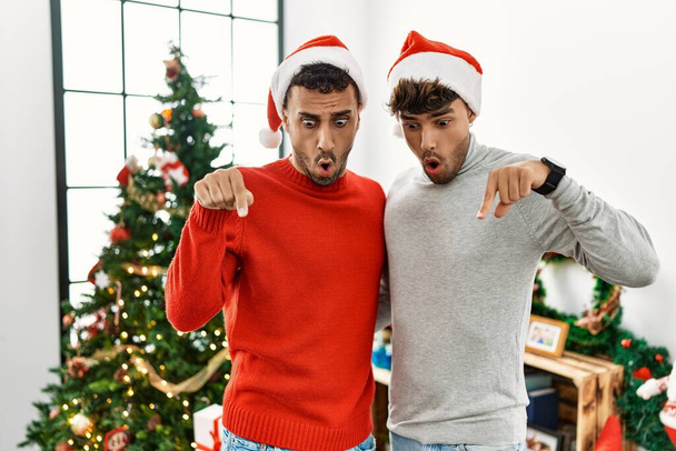Young gay couple standing by christmas tree wearing hat pointing down with fingers showing advertisement, surprised face and open mouth  - Photo, Image