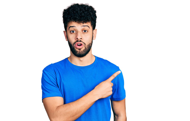 Fiatal arab férfi szakáll visel alkalmi kék póló meglepett mutatva ujjal oldalra, nyitott szájú elképedt kifejezés.  - Fotó, kép