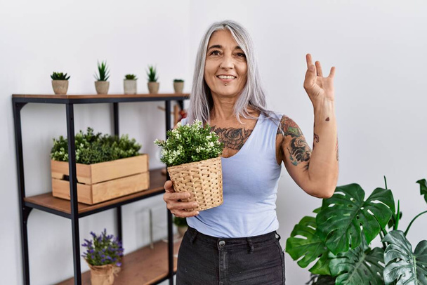 Middle age grey-haired woman holding green plant pot at home smiling positive doing ok sign with hand and fingers. successful expression.  - Valokuva, kuva