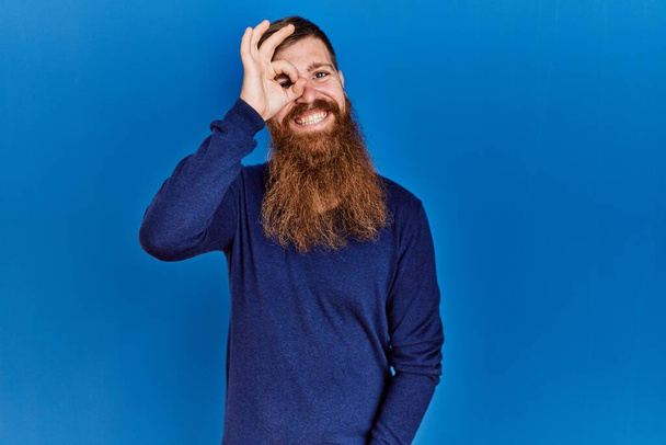 Redhead man with long beard wearing casual blue sweater over blue background doing ok gesture with hand smiling, eye looking through fingers with happy face.  - Foto, imagen