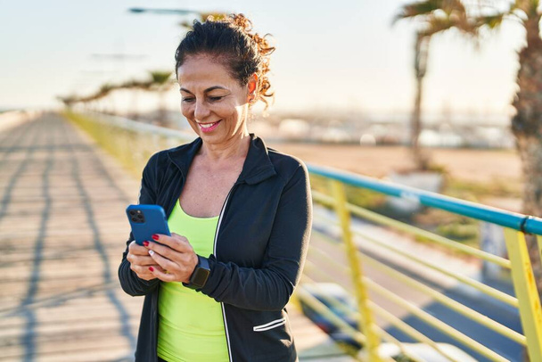Middle age hispanic woman working out with smartphone at promenade - 写真・画像