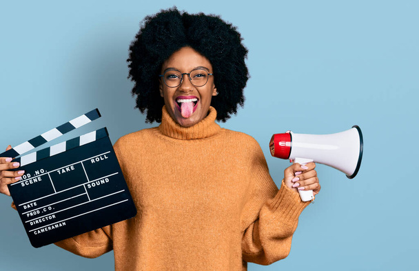 Young african american woman holding video film clapboard and megaphone sticking tongue out happy with funny expression.  - Photo, Image