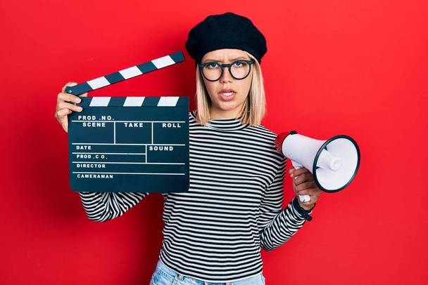 Beautiful blonde woman holding video film clapboard and megaphone clueless and confused expression. doubt concept.  - Photo, Image