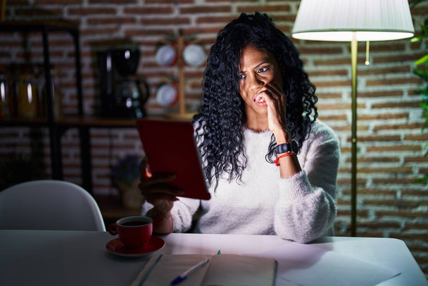 Middle age hispanic woman using touchpad sitting on the table at night looking stressed and nervous with hands on mouth biting nails. anxiety problem.  - Photo, Image
