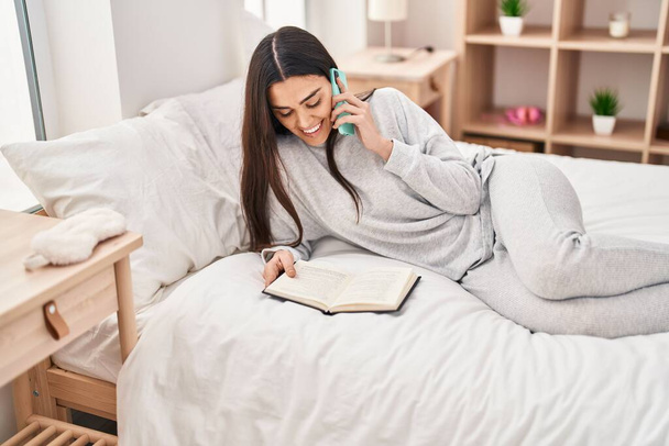 Молода іспаномовна жінка розмовляє на смартфоні читаючи книгу в спальні
 - Фото, зображення
