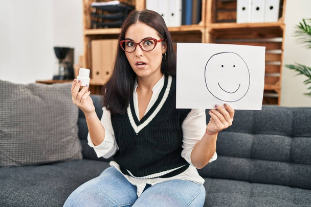Junge hispanische Frau, die an Depressionen arbeitet, hält glückliche Emotionen auf Papier und Tabletten im Schockgesicht, sieht skeptisch und sarkastisch aus, überrascht mit offenem Mund  - Foto, Bild