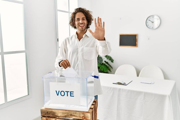 Giovane uomo ispanico che vota mettendo busta nelle urne rinunciando a dire ciao felice e sorridente, amichevole gesto di benvenuto  - Foto, immagini
