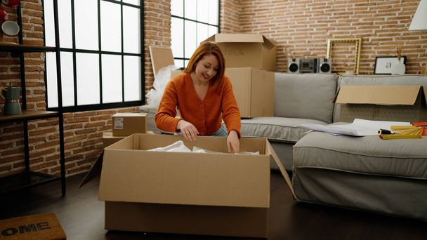 Young redhead woman smiling confident unpacking cardboard box at new home - Photo, Image
