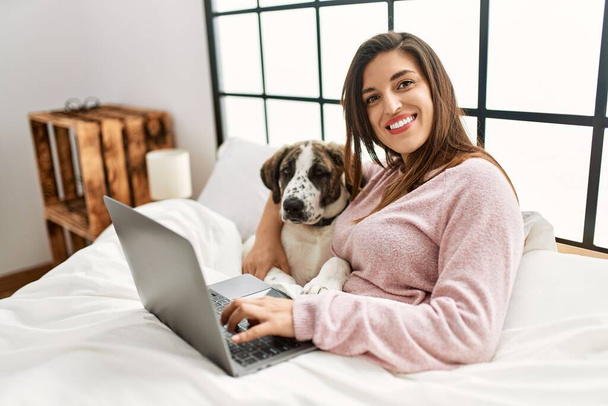 Młoda kobieta za pomocą laptopa przytulanie pies siedzi na łóżku w sypialni - Zdjęcie, obraz