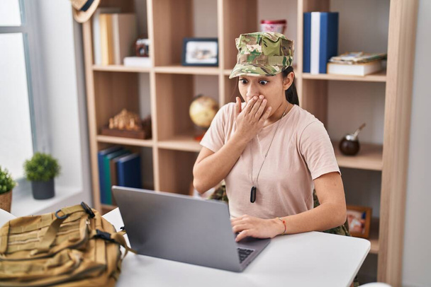 Nuori eteläaasialainen nainen yllään naamiointi armeijan univormu tekee videopuhelu kattaa suun käsin, järkyttynyt ja pelkää erehdystä. yllättynyt ilme  - Valokuva, kuva