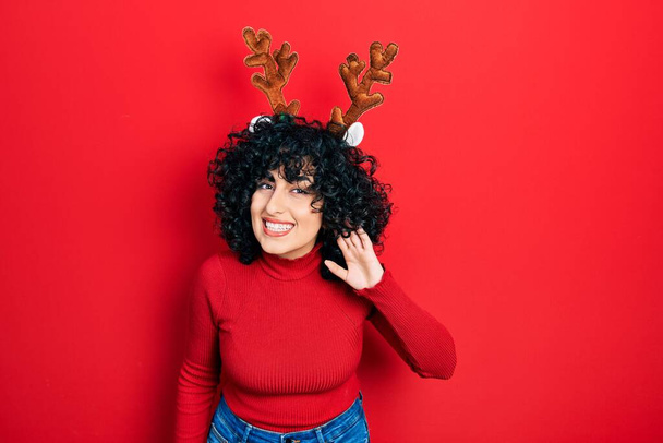 Young middle east woman wearing cute christmas reindeer horns smiling with hand over ear listening an hearing to rumor or gossip. deafness concept.  - Foto, Imagem