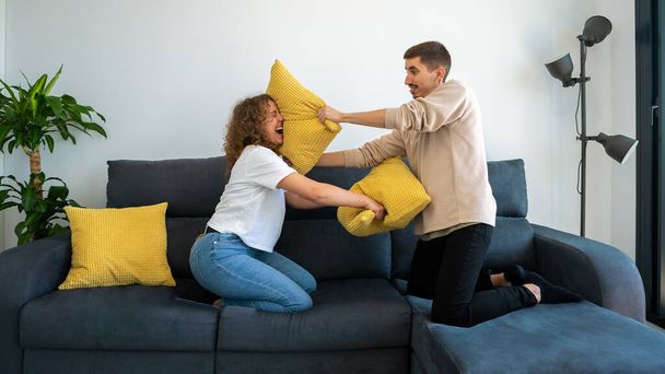 Couple of two happy boyfriends having fun playing together on the sofa at home fighting with cushion enjoying. Pillows war indoors at the morning - Foto, Imagen