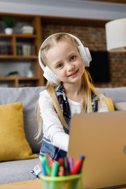 Portrait of cute primary school girl studying at home using laptop computer. Smiling child wearing white earphones working on computer. Distance learning. Children home education. - 写真・画像