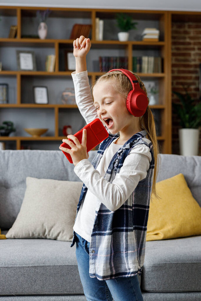 Happy little girl wearing red headphones dancing and singing using smartphone like microphone. Cute child enjoy listening favorite music tracks in mobile application Home leisure, happy childhood. - 写真・画像