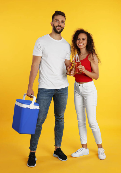 Happy couple with cool box and bottles of beer on yellow background - 写真・画像