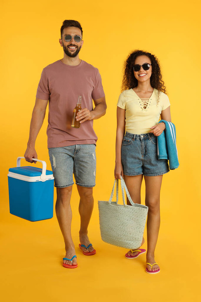 Happy young couple with cool box, bottle of beer and beach accessories on yellow background - Foto, afbeelding