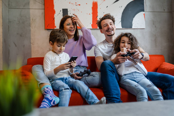 group of people caucasian family man and woman mother and father with children boy and girl sitting on the sofa at home playing video game console using joystick or controller real people concept - Photo, Image