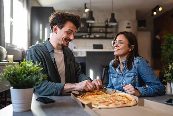 Genç beyaz erkek ve kadın modern karı-koca evde pizza yiyorlar. Erkek ve kadın arkadaşlar birlikte yemek yiyorlar. Ev hayatı kavramı ile zaman geçiriyorlar. - Fotoğraf, Görsel