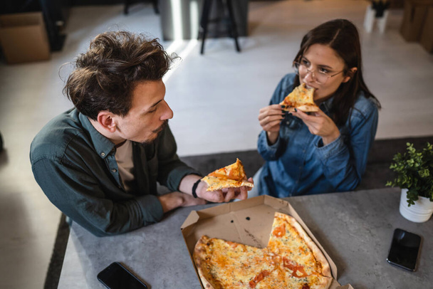 Genç beyaz erkek ve kadın modern karı-koca evde pizza yiyorlar. Erkek ve kadın arkadaşlar birlikte yemek yiyorlar. Ev hayatı kavramı ile zaman geçiriyorlar. - Fotoğraf, Görsel