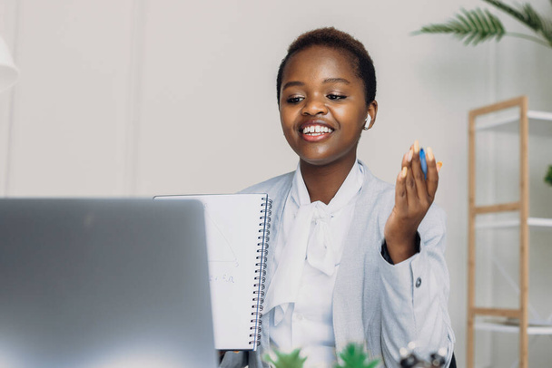 Afro amerykańska kobieta nosić słuchawki bezprzewodowe siedzieć przy biurku przez laptopa mówiąc i zadając pytania. Technologia biznesowa. Kobieta pracująca na laptopie. Kształcenie na odległość. - Zdjęcie, obraz