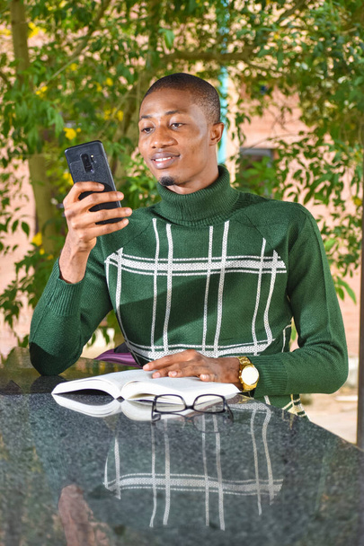 A light skinned african guy holding a smart phone and wearing a turtle-neck polo sits with a book in front of him during leisure time - Fotografie, Obrázek