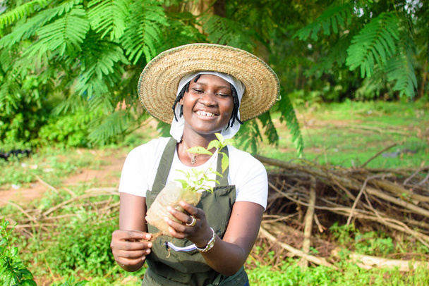 Happy African female gardener, florist or horticulturist wearing an apron and a hat, holding a bag of plant as she works in a green and colorful flowers garden - Foto, immagini