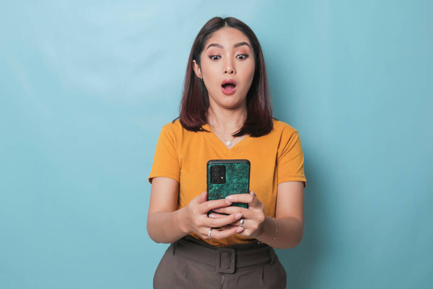 Surprised Asian woman looking at her smartphone over blue background - Zdjęcie, obraz