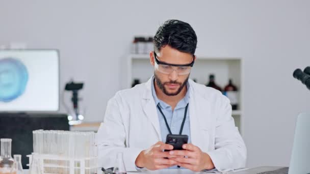 Happy scientist researching statistics and medical journals on phone, wearing safety glasses and a lab coat. Young male chemist or medical researcher checking data and looking relaxed in laboratory. - Filmmaterial, Video