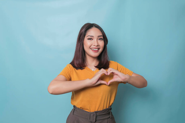 Attractive young Asian woman feels happy and romantic shapes heart gesture expresses tender feelings wears casual yellow t-shirt against blue background. People affection and care concept - Photo, image