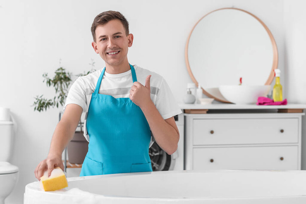 Young man cleaning bathtub with sponge and showing thumb-up in room - Foto, imagen