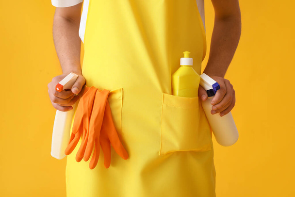 Young man in apron with cleaning supplies on yellow background, closeup - Valokuva, kuva