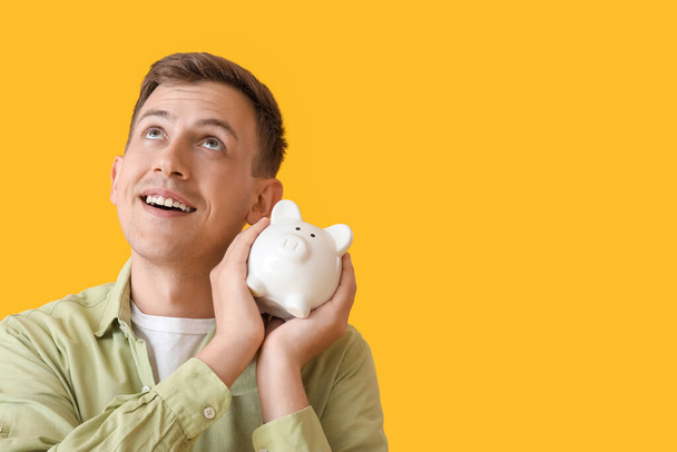 Young man with white piggy bank on yellow background - Fotoğraf, Görsel
