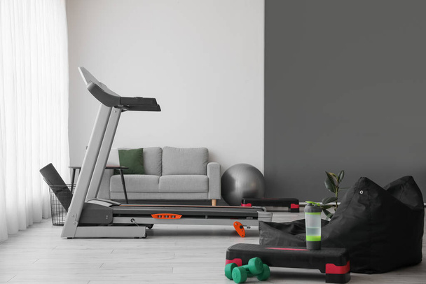 Interior of light living room with treadmill, sofa and sport equipment - Foto, afbeelding