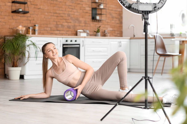 Young woman with foam roller recording video in kitchen - Foto, afbeelding