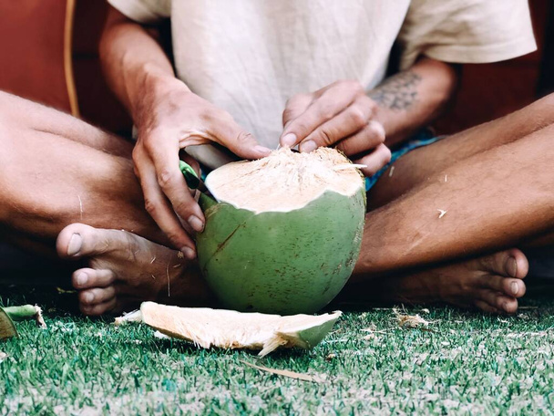 A close-up of a person opening a fresh coconut with a knife while sitting cross-legged on the grass - Fotografie, Obrázek