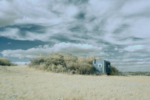 An infrared shot of dry bushes near an old blue trailer in the yellow meadow under the blue sky and white clouds - Foto, afbeelding