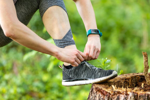 A closeup shot of a Young woman makes fitness and ties shoelaces in nature with a smartwatch on her arm - 写真・画像