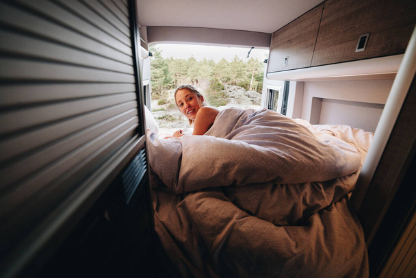 Młoda naga kobieta leżąca na łóżku pod kocem i patrząca przez ramię na malowniczy las iglasty za otwartymi drzwiami kampera podczas podróży latem - Zdjęcie, obraz