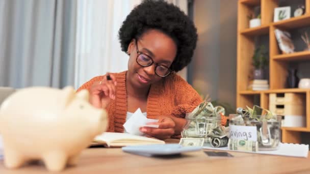 Financiën, budget en geld besparen door geld te storten in een spaarvarken bank na het berekenen van een budget in een huiskamer. Glimlachende, gelukkige en vrolijke vrouw met afro planning van haar uitgaven en uitgaven. - Video