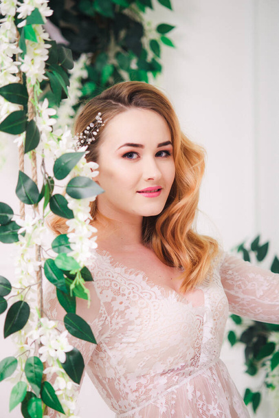 Young attractive bride with blond hair with wedding makeup and hairstyle in a white lace peignoir in a bright interior - Foto, Imagem
