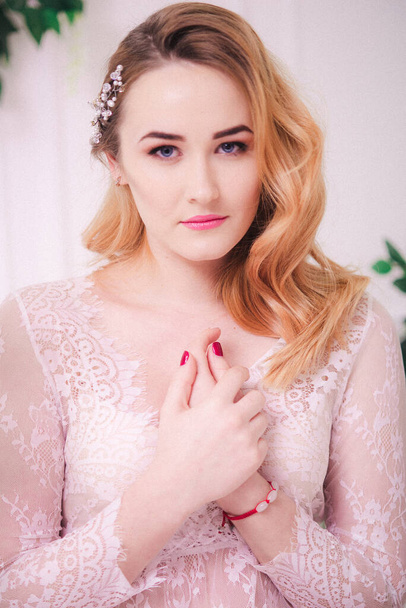 Young attractive bride with blond hair with wedding makeup and hairstyle in a white lace peignoir in a bright interior - Photo, image