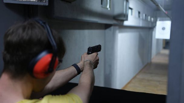 Close-up of man shooter in tactical headphones aiming pistol at target in shooting range. Shooting practicing with gun in rifle range. Extreme hobby concept - 写真・画像