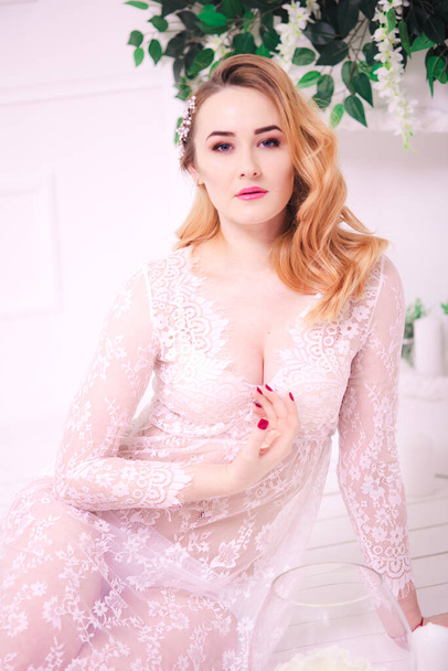 Young attractive bride with blond hair with wedding makeup and hairstyle in a white lace peignoir in a bright interior - Foto, imagen