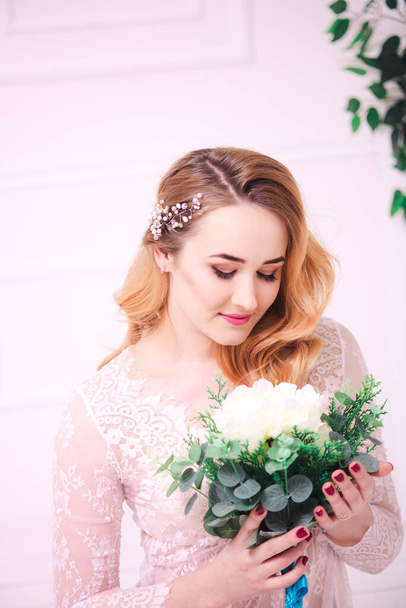 Young attractive bride with blond hair with wedding makeup and hairstyle in a white lace peignoir with bride's bouquet in a bright interior - Foto, Imagem