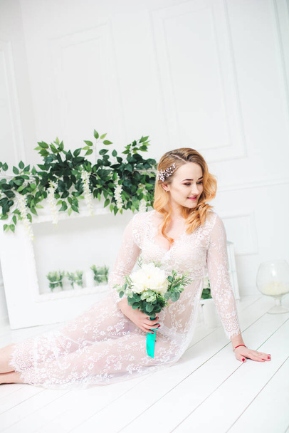 Young attractive bride with blond hair with wedding makeup and hairstyle in a white lace peignoir with bride's bouquet in a bright interior - Foto, immagini