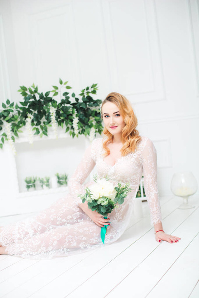 Young attractive bride with blond hair with wedding makeup and hairstyle in a white lace peignoir with bride's bouquet in a bright interior - Фото, зображення