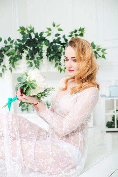 Young attractive bride with blond hair with wedding makeup and hairstyle in a white lace peignoir with bride's bouquet in a bright interior - Foto, Imagen